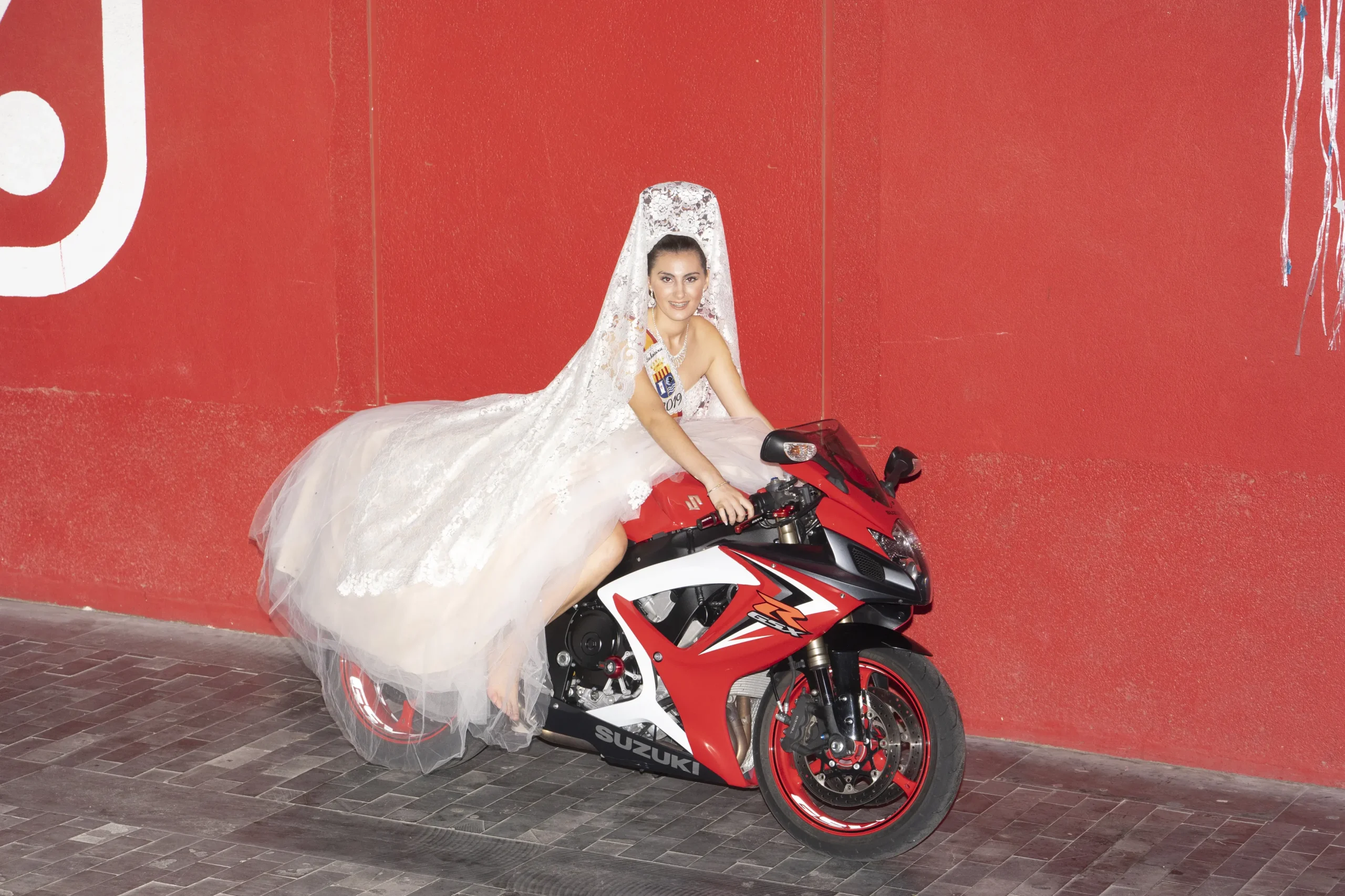 Retrato de una joven vestida con traje regional de gala y mantilla sentada sobre una motocicleta Suzuki roja, perteneciente a la serie "Educandos de Benejúzar" de Carlos Aguilera.