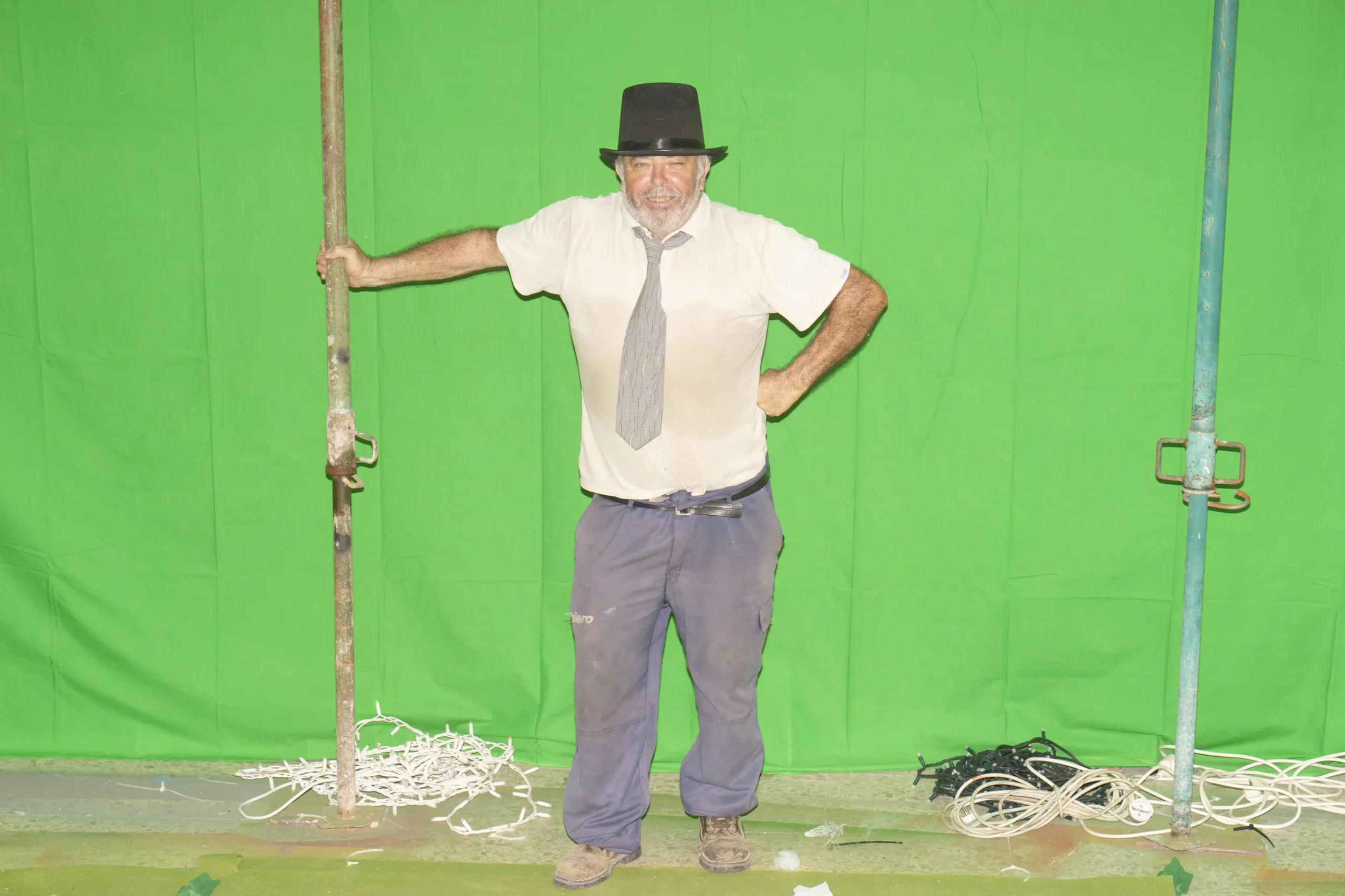 Retrato de un hombre mayor con ropa de trabajo posando con una chistera negra y corbata ante un fondo de croma verde y andamios, de la serie "Educandos de Benejúzar" de Carlos Aguilera.