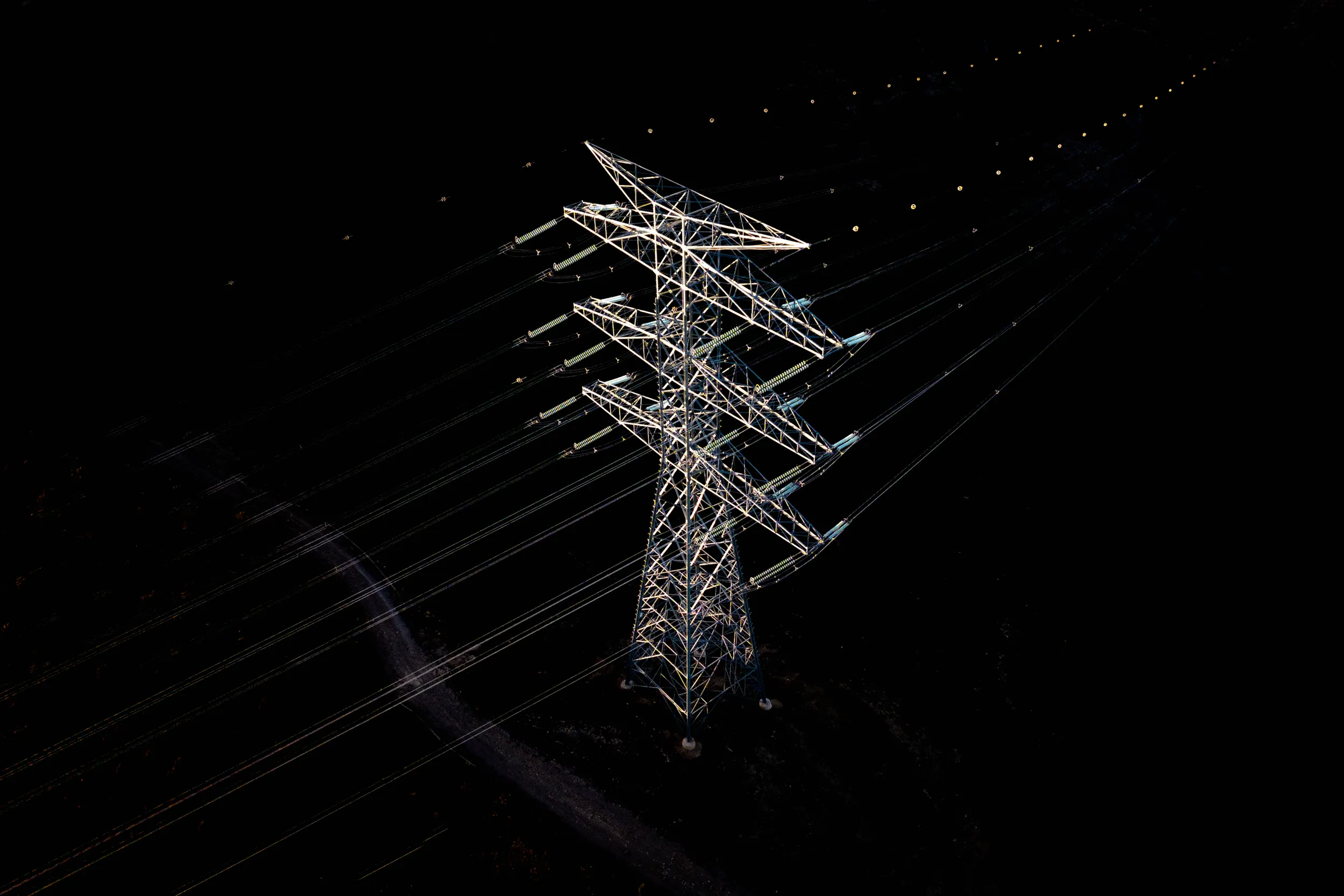 Vista aérea nocturna de una torre de alta tensión y su cableado iluminados sobre un fondo negro, obra de Andreu Esteban en Terra Nullius.