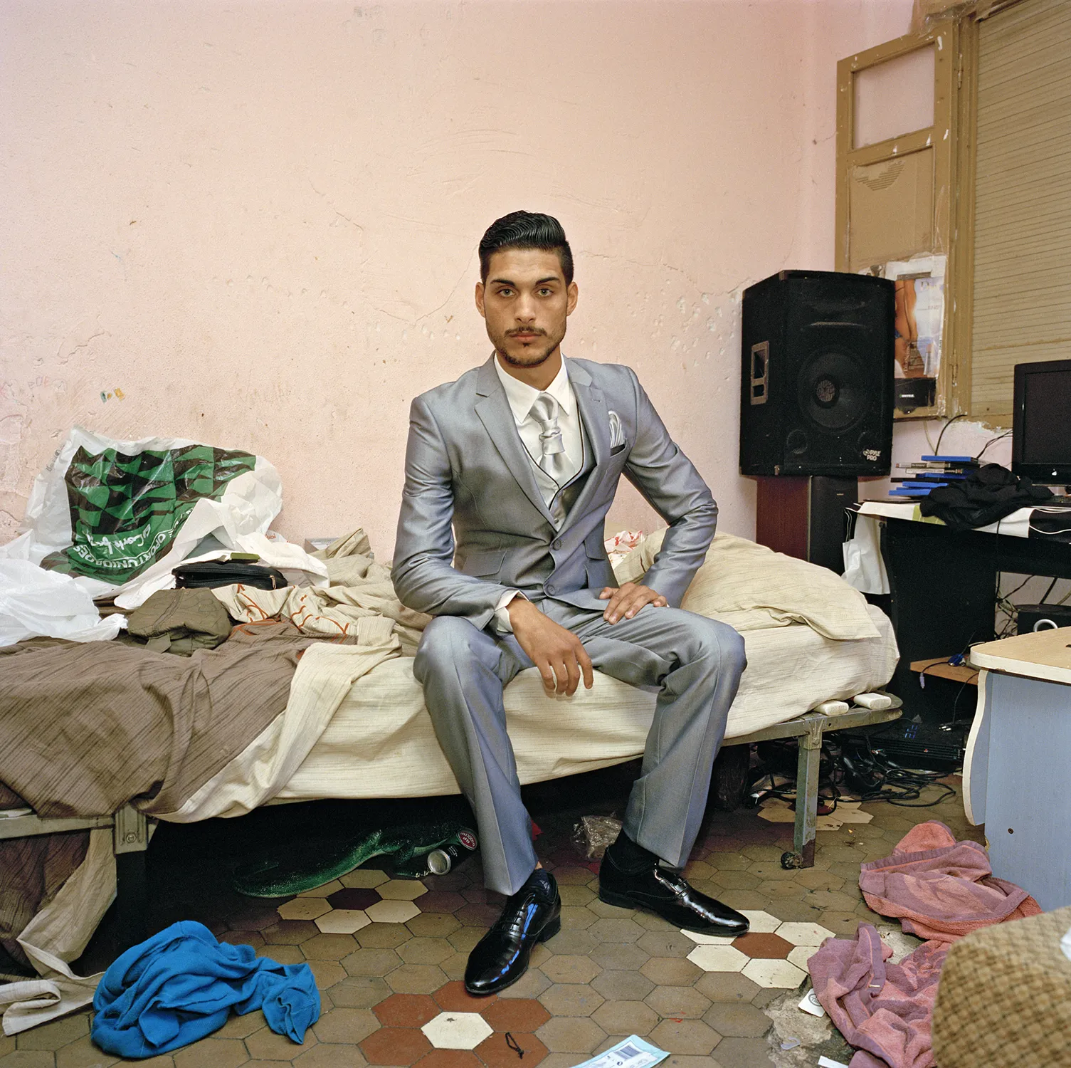 Hombre joven vistiendo un traje gris brillante sentado sobre una cama en una habitación desordenada, perteneciente al proyecto Quisiera que siempre fuera así de Laura Silleras.