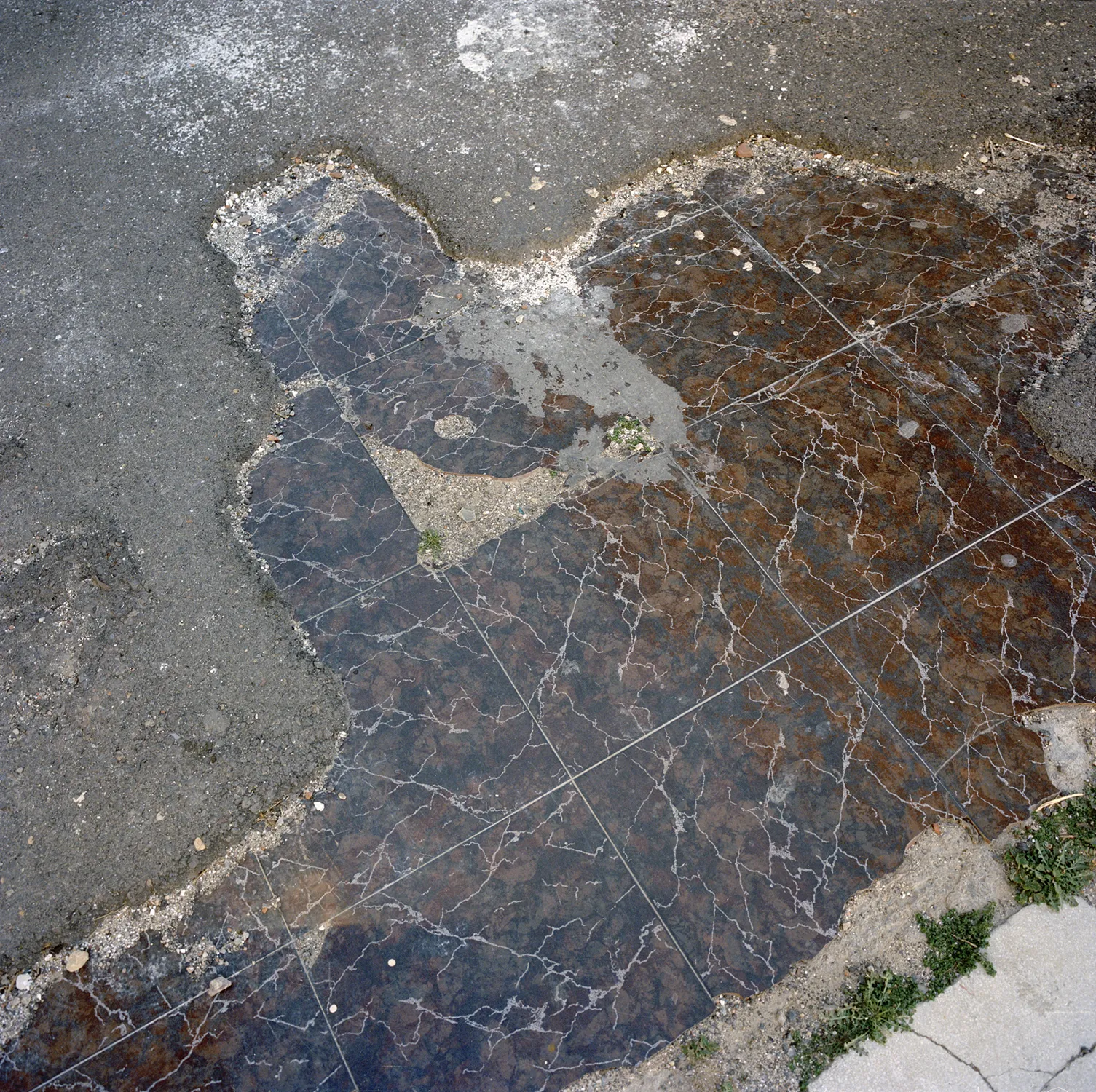 Detalle de un suelo donde unas baldosas oscuras y rotas se mezclan directamente con el asfalto de la calle, del proyecto Quisiera que siempre fuera así de Laura Silleras.