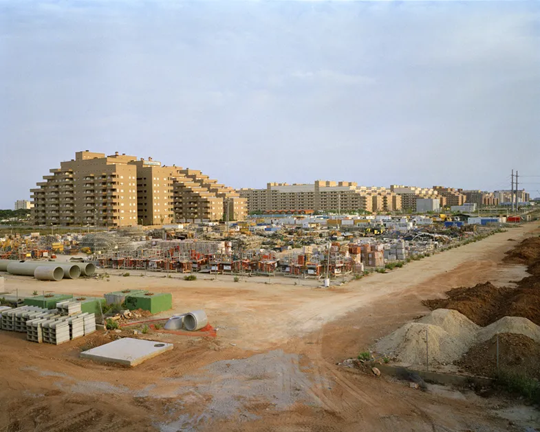 Explanada de tierra llena de materiales de construcción frente a grandes bloques de apartamentos escalonados, obra del proyecto La idea de éxito de Markel Redondo.
