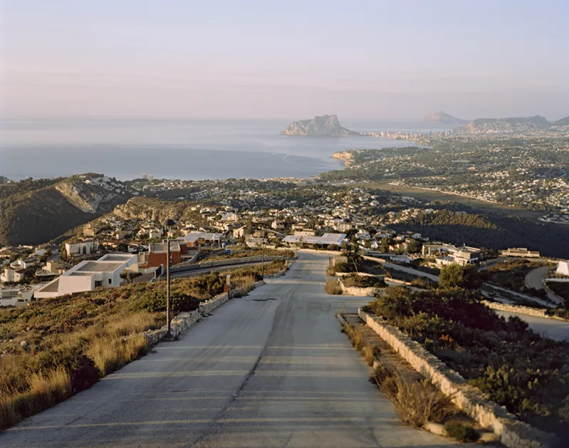 Carretera asfaltada que desciende hacia una gran urbanización costera con el Peñón de Ifach al fondo, del proyecto La idea de éxito de Markel Redondo.